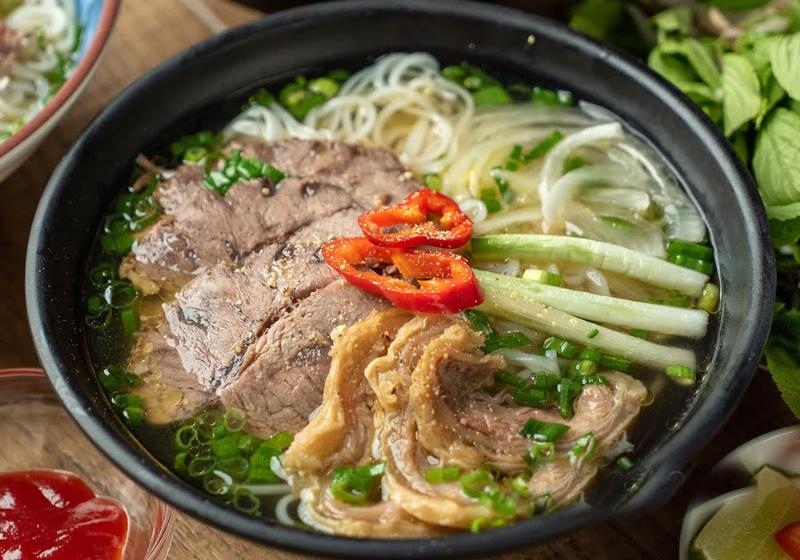 Một tô phở Việt Nam với đầy đủ thịt bò, hành lá, rau thơm, và nước dùng trong veo, bốc khói nghi ngút